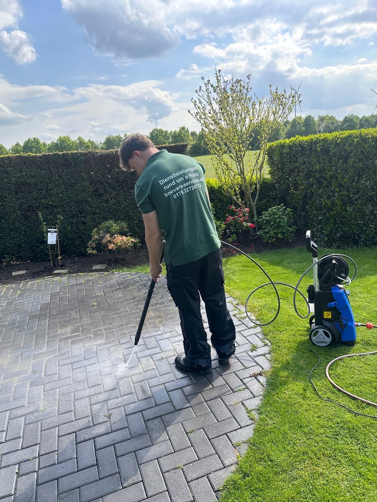 Man in green shirt pressure washing a brick patio with a blue power washer and hose in a sunny backyard.